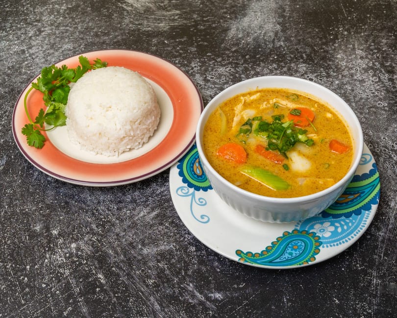 A bowl of yellow curry with vegetables and chicken next to a plate of white rice and cilantro.
