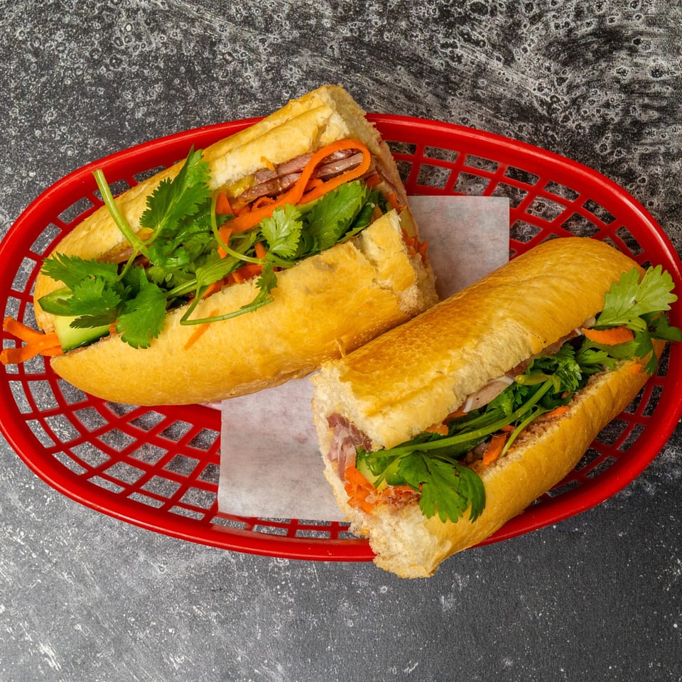 Two banh mi sandwiches with meat, carrots, and cilantro in a red plastic basket.