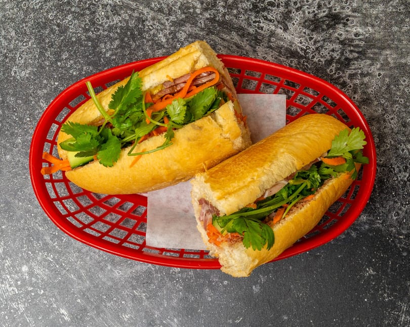 Two banh mi sandwiches with meat, carrots, and cilantro in a red basket on a dark background.