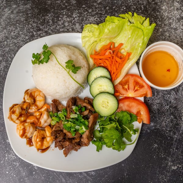 A white plate with rice, shrimp, beef, lettuce, carrots, cucumbers, tomatoes, and a side of sauce.