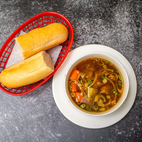 A bowl of beef stew with carrots and green onions, served with two baguettes in a red basket.
