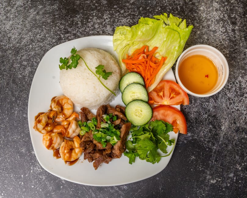A white plate with rice, shrimp, beef, lettuce, carrots, cucumbers, tomatoes, and a side of sauce.