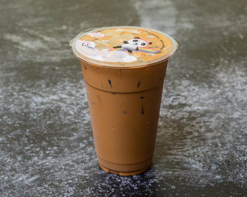 A tall plastic cup of iced brown bubble tea with a panda on the lid sits on a dark surface.