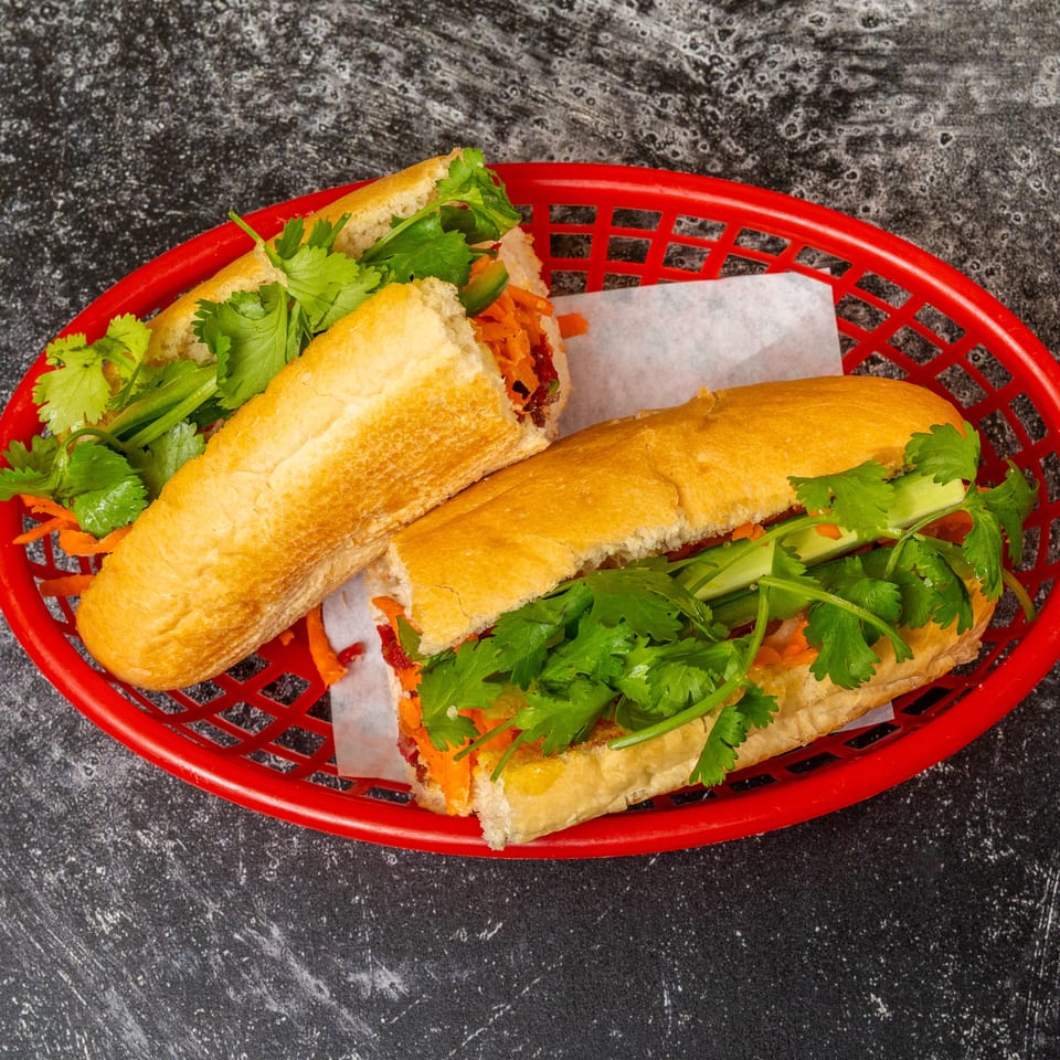 Two banh mi sandwiches with fresh cilantro, carrots, and cucumbers in a red basket.
