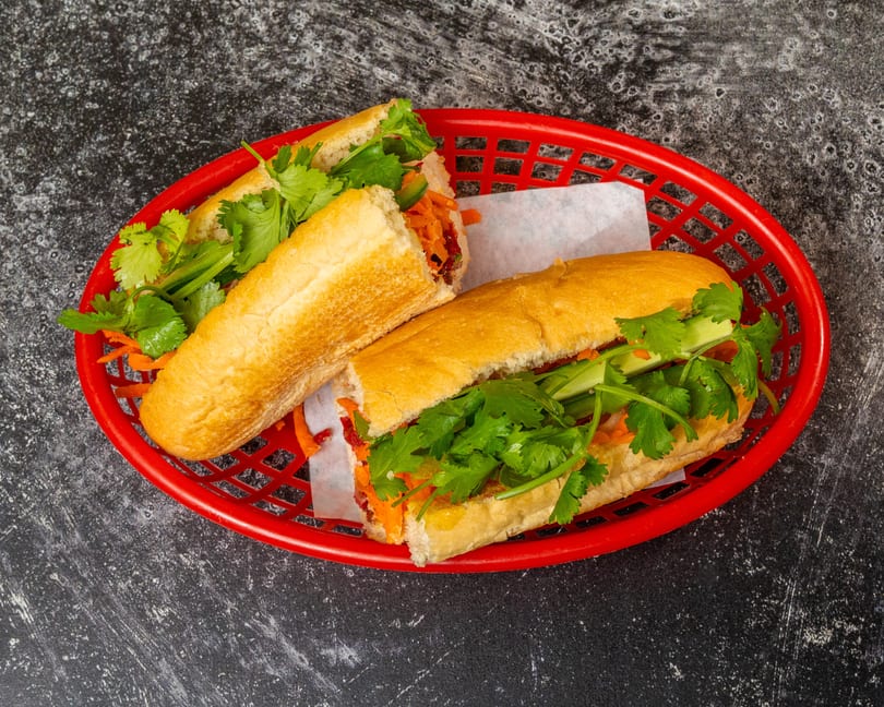 Two banh mi sandwiches with fresh cilantro, carrots, and cucumbers in a red basket.