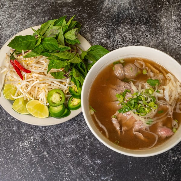 A bowl of pho with beef, noodles, and broth, alongside a plate of fresh herbs and garnishes.