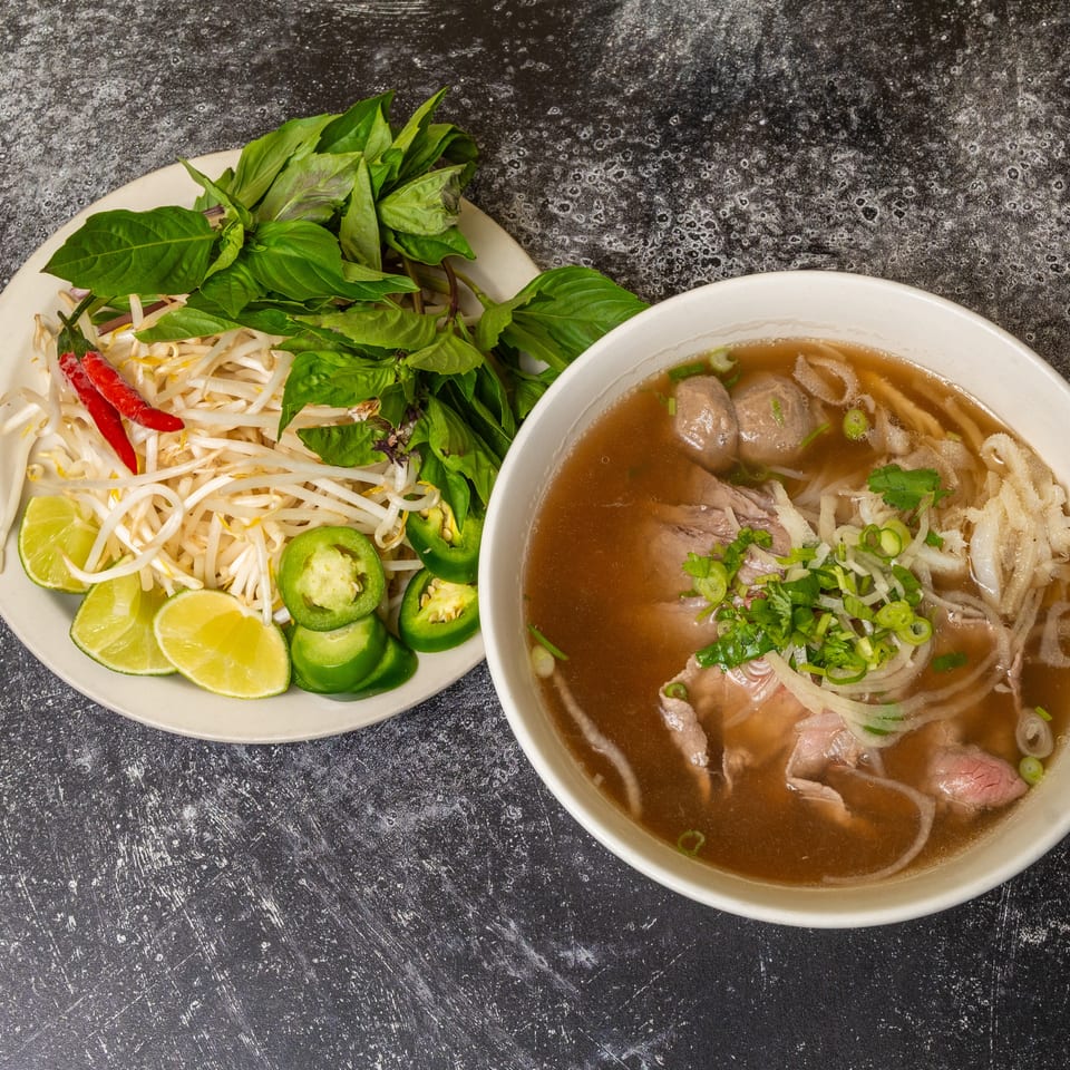 A bowl of pho with beef, noodles, and broth, alongside a plate of fresh herbs and garnishes.