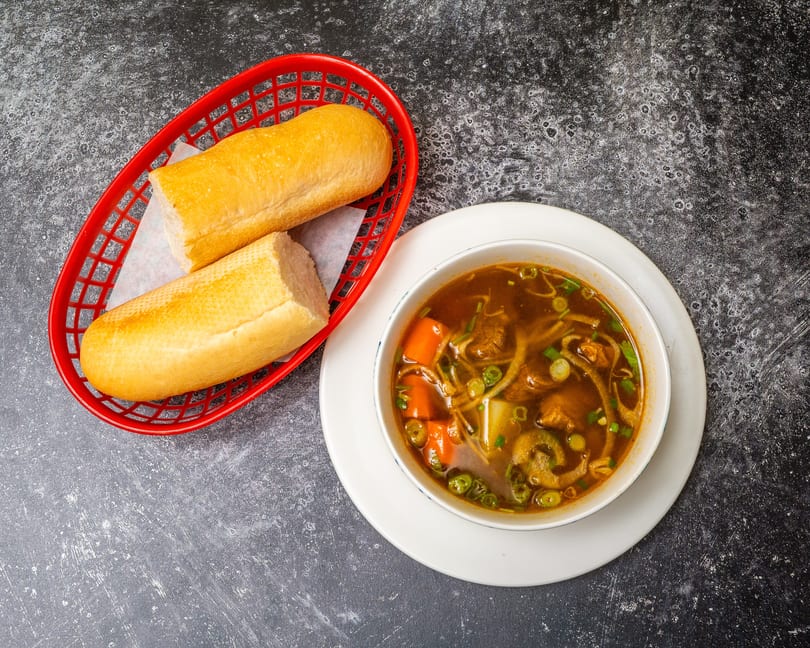 A bowl of beef stew with carrots and green onions, served with two baguette halves in a red basket.