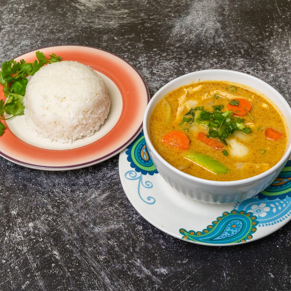 A bowl of yellow curry with chicken and vegetables, served with a side of white rice and cilantro.