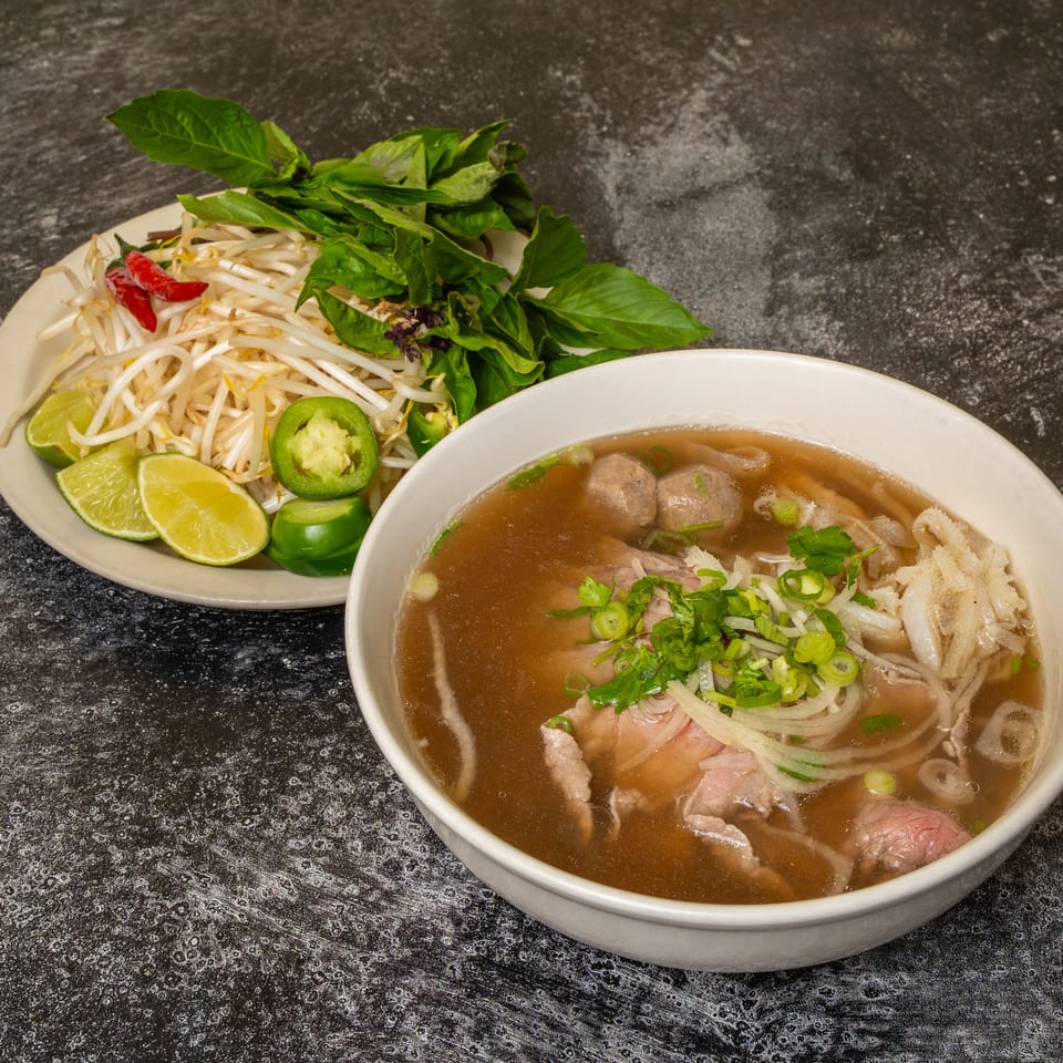 A bowl of pho with beef, noodles, and broth, served with a side of bean sprouts, basil, limes, and jalapeños.
