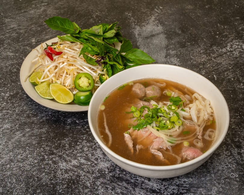 A bowl of pho with beef, noodles, and broth, served with a side of bean sprouts, basil, lime, and jalapeños.