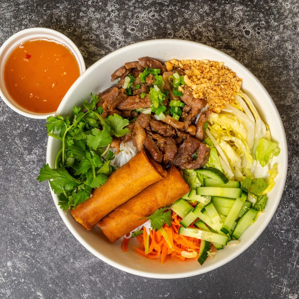 Vietnamese vermicelli bowl with grilled beef, crispy spring rolls, fresh vegetables, and a side of sauce.