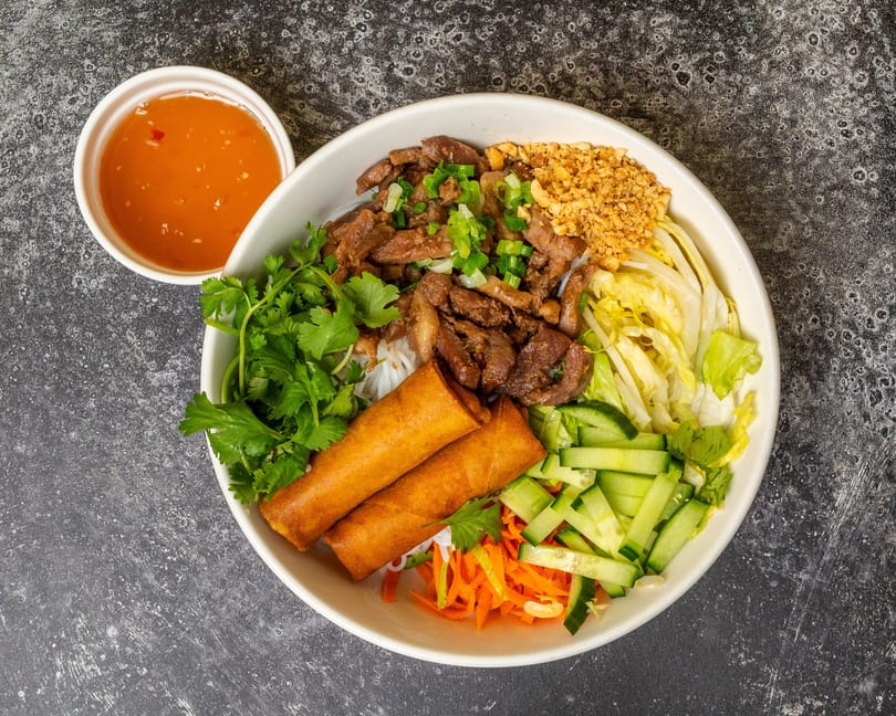 A bowl of Vietnamese vermicelli with grilled pork, spring rolls, vegetables, and a side of sauce.
