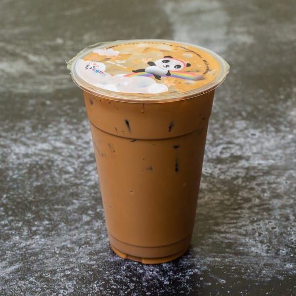 A large plastic cup of iced Thai tea with a panda-themed lid sits on a dark, speckled surface.